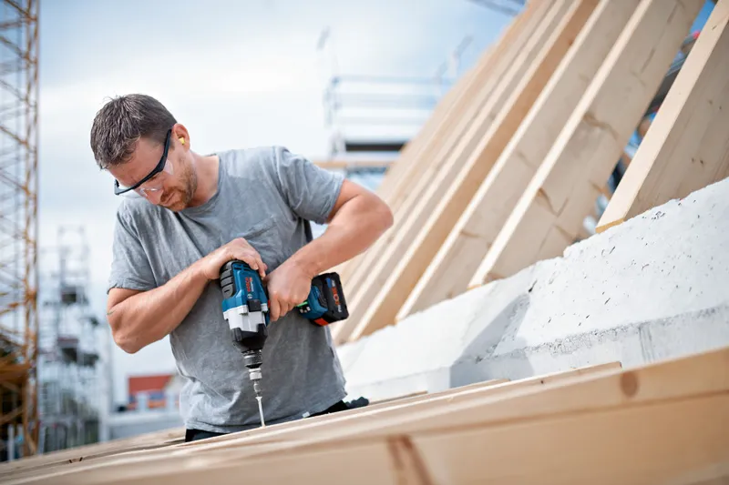 GDS 18V-1000 Person wearing safety equipment uses a cordless impact wrench to drive a screw into wood.