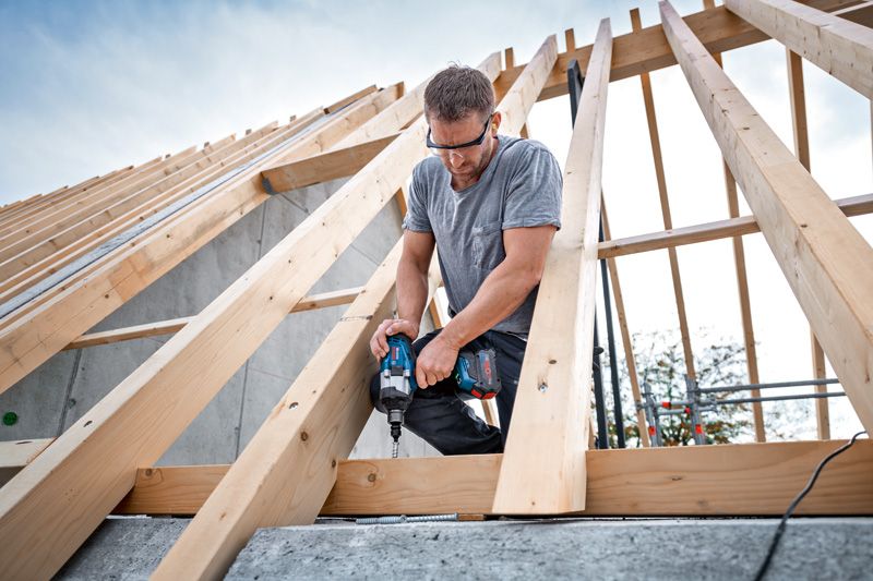 A person wearing safety equipment uses a cordless impact wrench to fasten wood beams.
