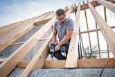 A person wearing safety equipment uses a cordless impact wrench to fasten wood beams.