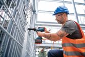 A person wearing safety equipment uses a cordless impact wrench to fasten a metal fence.
