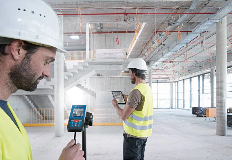 Two people wearing safety equipment measure distance in a large building using a laser measure.