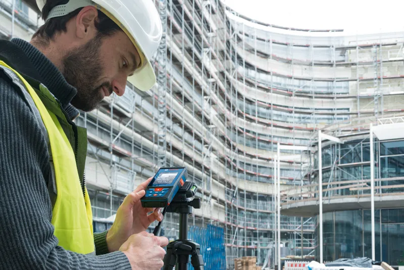 A person wearing safety equipment sets up a laser measure on a tripod at a construction site.