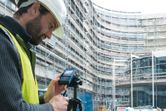 A person wearing safety equipment operates a laser measure at a construction site.