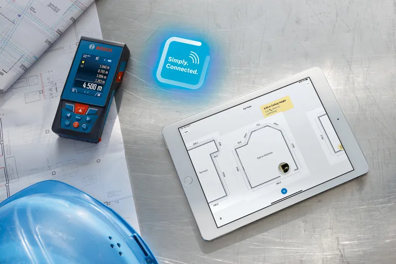 Laser measure next to construction plans, a tablet with floor layout, and a blue safety helmet.