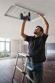 A person wearing safety equipment installs a ceiling light fixture using a power drill.