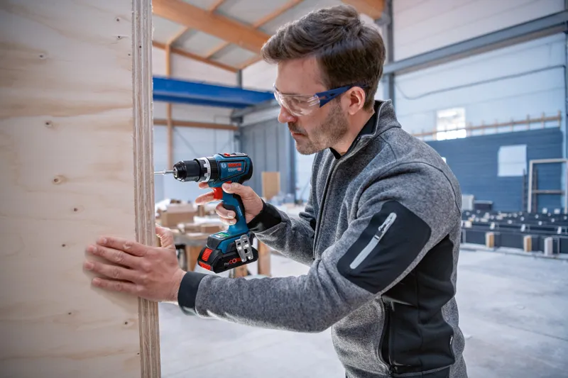 GSB 18V-90 C Person wearing safety equipment drills into a wooden board inside a workshop.