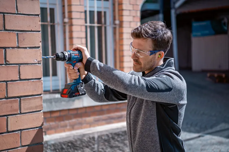 GSB 18V-90 C A person wearing safety equipment drills into a brick wall with a cordless power drill.