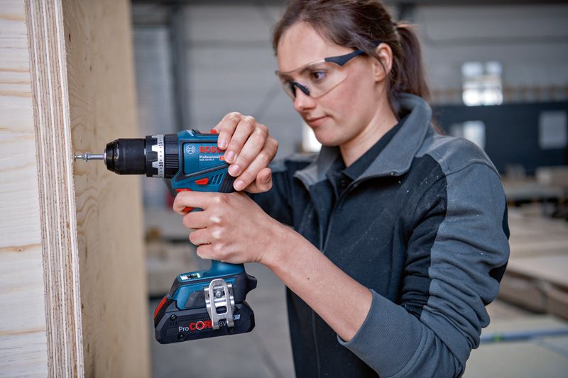 GSB 18V-90 C A person wearing safety equipment uses a cordless drill on a wooden panel.