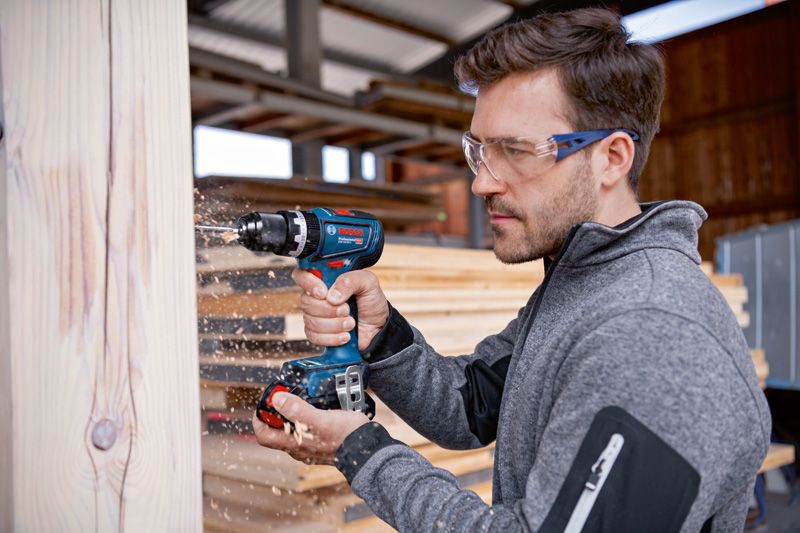 GSB 18V-90 C A person wearing safety equipment drills into a wooden beam in a workshop.