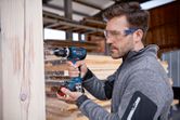 A person wearing safety equipment drills into a wooden beam in a workshop.