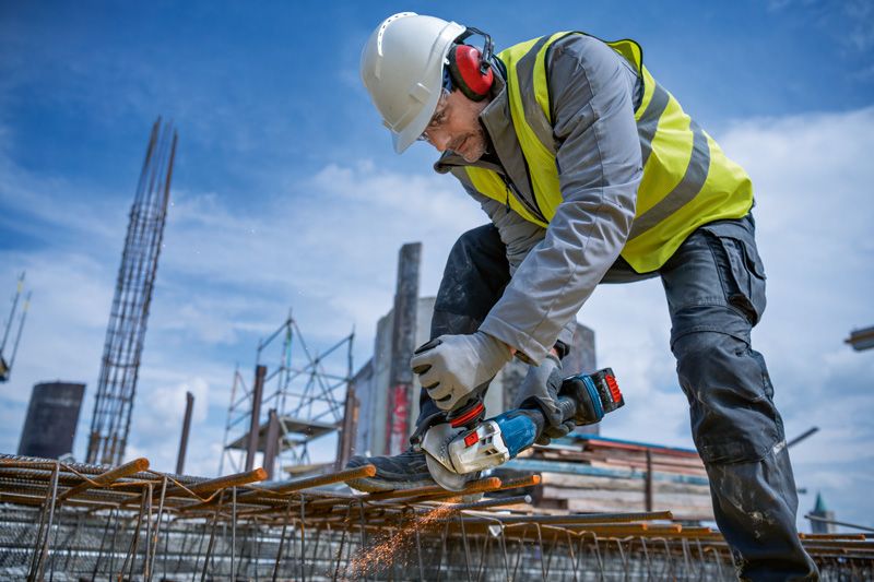 A person wearing safety equipment grinds rebar with a cordless angle grinder on a construction site.
