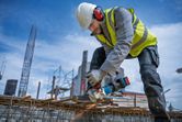 A person wearing safety equipment grinds rebar with a cordless angle grinder on a construction site.