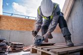 A person wearing safety equipment cuts tile on a pallet using a cordless angle grinder.