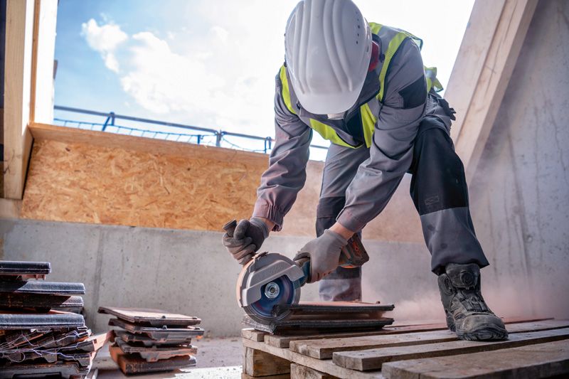 A person wearing safety equipment cuts tiles with a cordless angle grinder on a pallet.