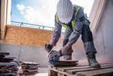 A person wearing safety equipment cuts tiles with a cordless angle grinder on a pallet.