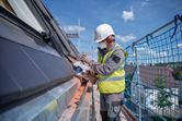 A person wearing safety equipment uses a cordless angle grinder on a building roof.