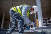 A person wearing safety equipment uses a cordless angle grinder to cut a concrete slab.