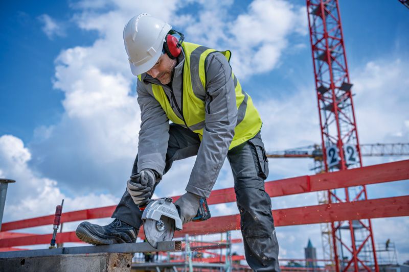 GWS 18V-180 P A person wearing safety equipment uses a cordless angle grinder to cut metal on a construction site.