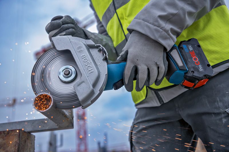 A person wearing safety equipment cuts a metal pipe with a cordless angle grinder.