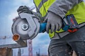 A person wearing safety equipment cuts metal pipe with a cordless angle grinder.