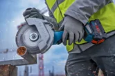 A person wearing safety equipment cuts a metal pipe with a cordless angle grinder.