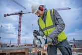 A person wearing safety equipment uses a cordless angle grinder to cut metal at a construction site.