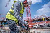 A person wearing safety equipment cuts rebar at a construction site with a cordless angle grinder.