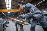 A worker wearing safety equipment grinds a metal beam in an industrial workshop.