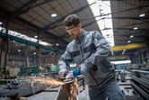 A person wearing safety equipment grinds metal with a cordless angle grinder in a factory.