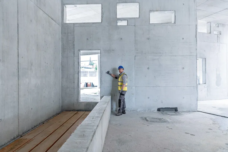 A person wearing safety equipment uses a laser measure against a concrete wall.