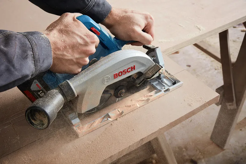 GKS 185-LI A person cuts a wooden board with a cordless circular saw, creating sawdust.