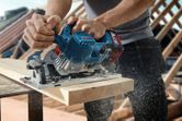 A person cuts a wooden board with a cordless circular saw, sawdust flying.