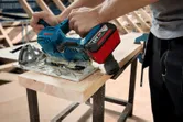 Person using a cordless circular saw to cut a wooden board on a workbench.