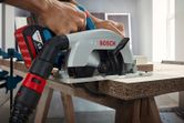 A person cuts a wooden board with a cordless circular saw attached to a vacuum hose.