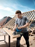 A person wearing safety equipment cuts wood with a cordless circular saw at a construction site.
