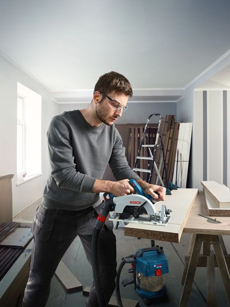 Person wearing safety equipment cuts wood with a cordless circular saw in a workshop.