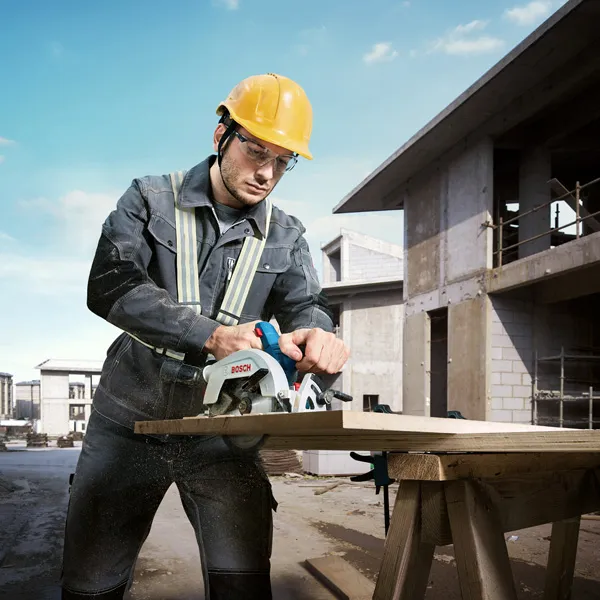 GKS 185-LI A person wearing safety equipment cuts wood with a cordless circular saw at a construction site.