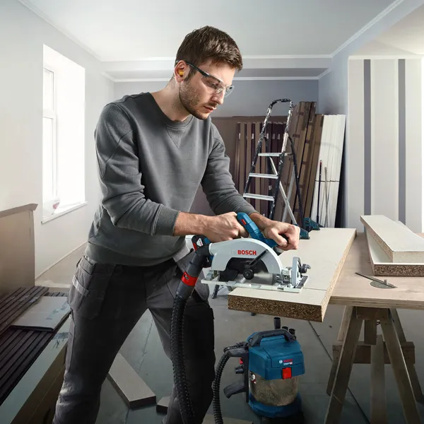 GKS 185-LI A person wearing safety equipment cuts wood with a cordless circular saw in a workshop.