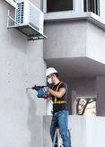 A person wearing safety equipment drills into a concrete wall beside an air conditioning unit.