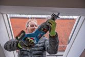 A person wearing safety equipment uses a cordless reciprocating saw to cut a window frame.