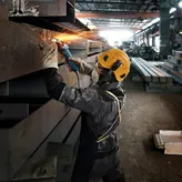 A person wearing safety equipment grinds a metal beam, creating bright sparks.