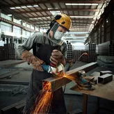 A person wearing safety equipment grinds a metal beam with an angle grinder, sparks flying.