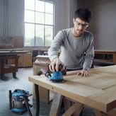 A person wearing safety equipment sands a wooden door with a connected orbital sander.