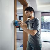 A person wearing safety equipment sands a wall with a cordless power sander.