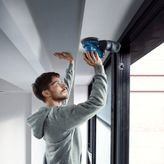 A person wearing safety equipment sands a ceiling edge using a power sander.