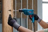 A person wearing safety equipment drills into a wooden board.