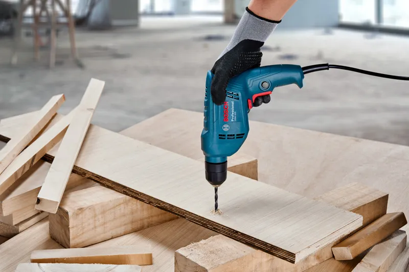 A person wearing safety equipment drills a hole in plywood on a workbench.