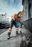 A person wearing safety equipment breaks pavement with a power breaker on a city street.