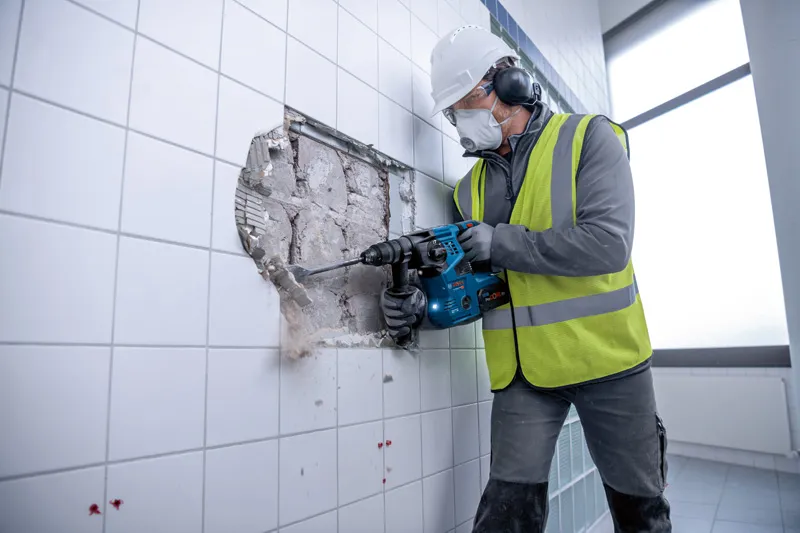 GBH 18V-28 CF Person wearing safety equipment uses a cordless rotary hammer to break through a tiled wall.
