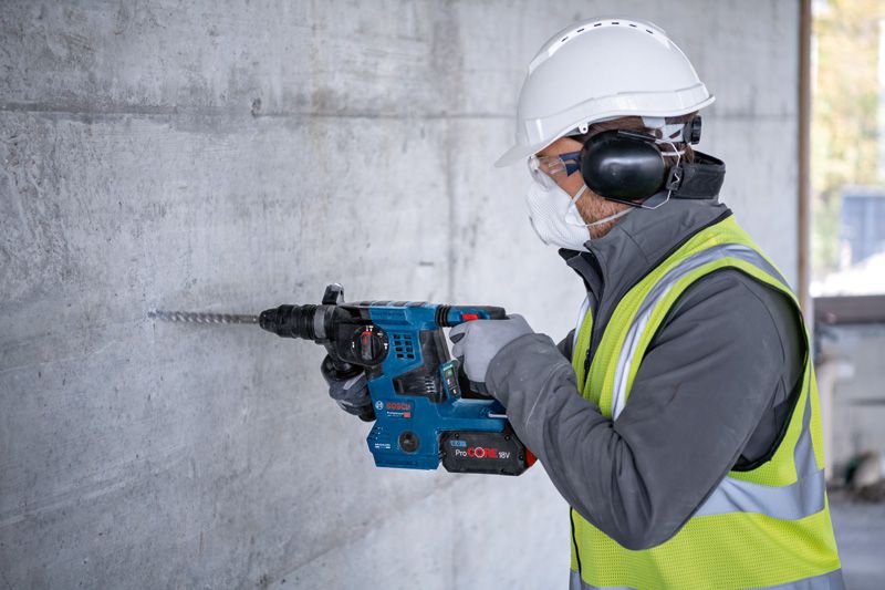 GBH 18V-28 CF A person wearing safety equipment drills into a concrete wall with a cordless rotary hammer.